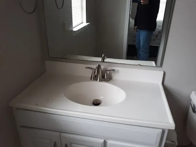 Bathroom vanity with white countertop, oval sink, and a silver faucet. A mirror reflects a person.