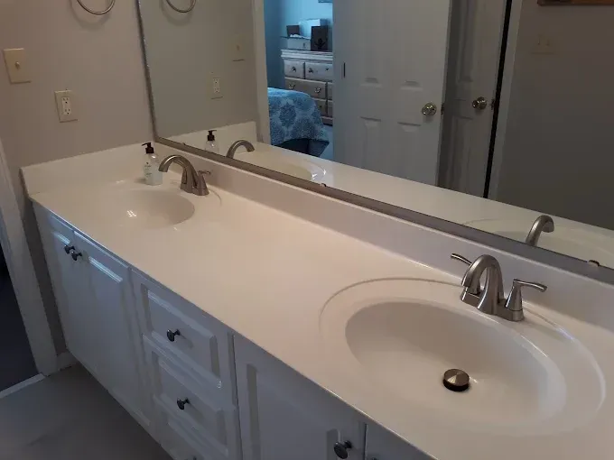 White double sink vanity in bathroom, with large mirror reflecting a bedroom.