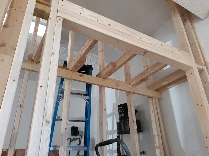 Wooden framing for an interior wall, including a header and studs, with a ladder and tools visible.