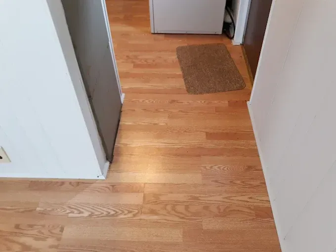 Hardwood-style flooring in a hallway with a small rug, two white walls, and a grey door.