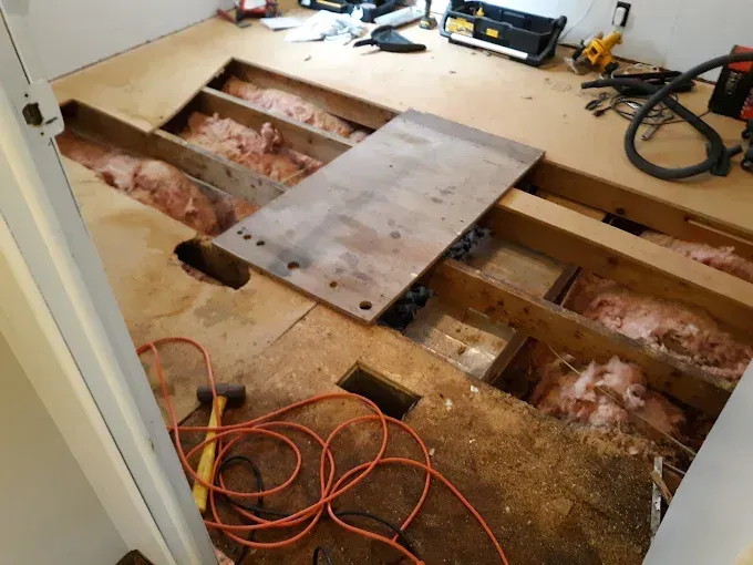 Room interior with exposed floor joists, insulation, and construction debris. Tools and a work light are present.