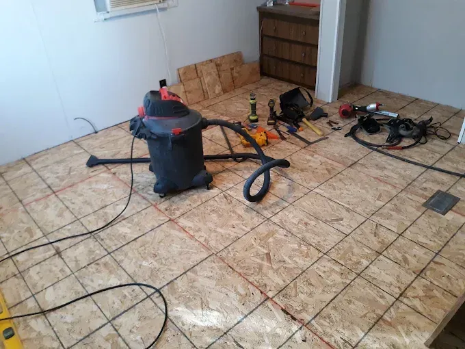 Room with exposed subfloor, tools, and a shop vac, ready for flooring installation.