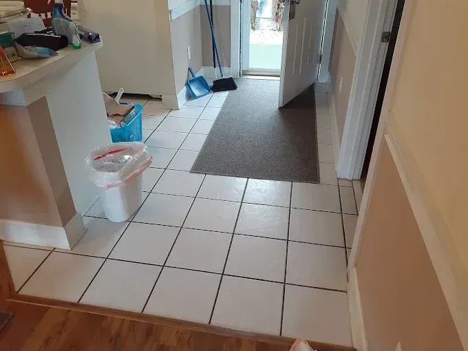 Tiled hallway leading to an open doorway with a doormat. A trash can and cleaning supplies are visible.