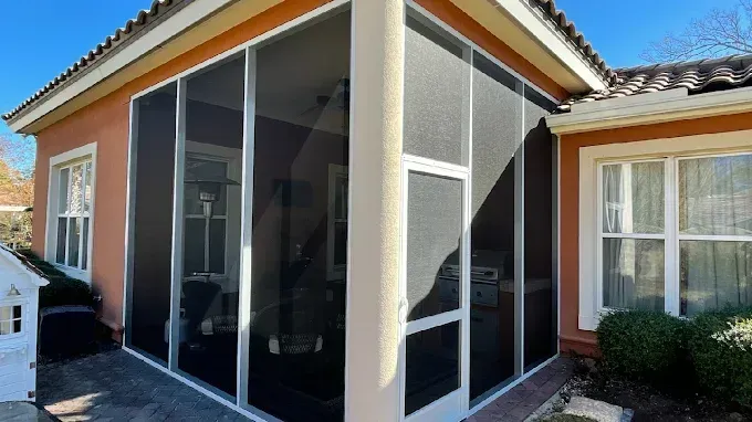 Screened-in porch attached to a peach-colored stucco house with white trim and a tiled roof, bright blue sky.