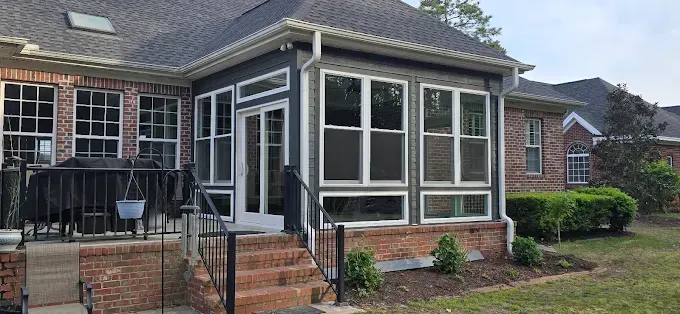 A screened-in porch with windows and a brick exterior. A yard is in the foreground.