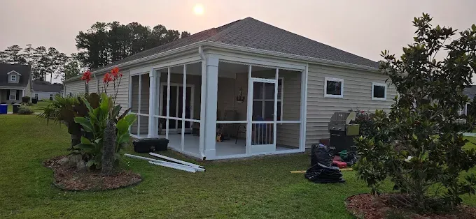 Backyard view of a house with a screened porch, grass, and plants. Overcast sky with sun visible.