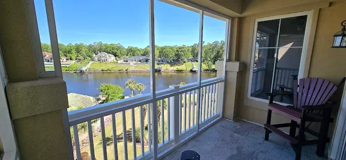 Balcony overlooking a lake with screen windows and a chair. Sunny day with trees and houses in the distance.