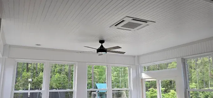 Bright sunroom interior, white ceiling with fan and AC unit. Large windows with outside view of green trees.