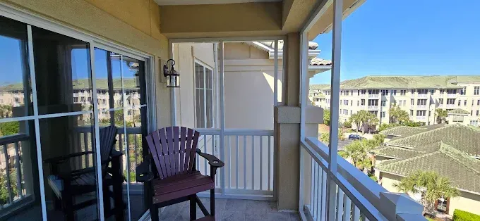 Balcony view: brown chair, sliding glass door, white railing, buildings with green roofs. Sunny sky.