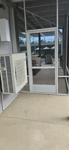 A screened-in porch with a wall-mounted air conditioner and a door opening to an outdoor area with patio furniture.