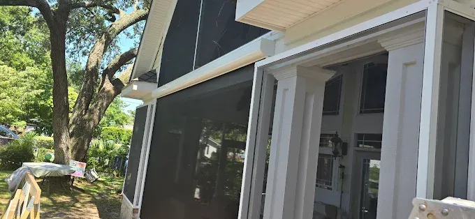 Screened-in porch with white trim, black screen, and large tree in the background. Bright sunny day.