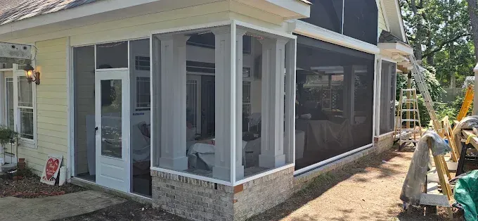 Screened porch with white trim, tan brick base, and dark screens. Yellow siding, trees in background.