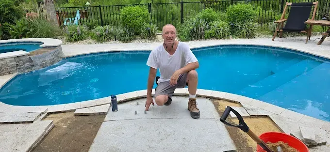Man kneels near pool, installing pavers. Blue water, green foliage, and tools visible.