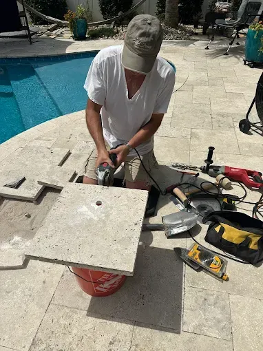 Man cutting a stone tile near a pool with power tools; outdoor setting.