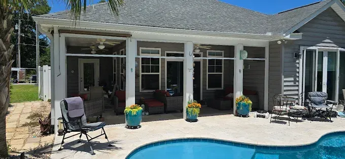 Backyard patio with pool. Beige patio tiles, light grey house with white columns, and a bright blue pool.