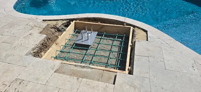 A square wooden form is set into the pool deck, containing rebar and a metal plate with mounting bolts.