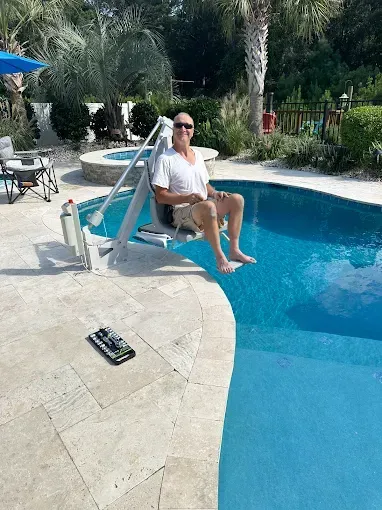 Man using a pool lift to enter a blue swimming pool on a sunny day.