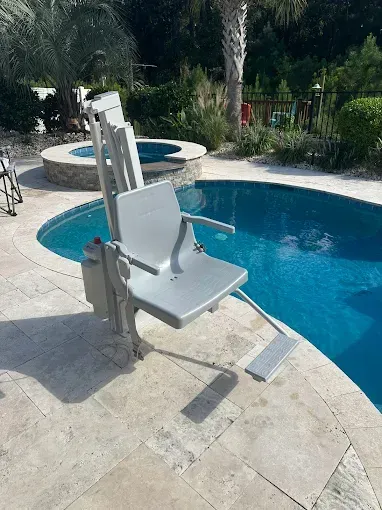 Pool lift next to a pool. A gray chair extends into the water. Surrounding stone deck.