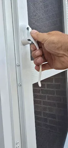 Hand pulling a white lever on a white screen door, revealing a gray screen, next to a brick wall.