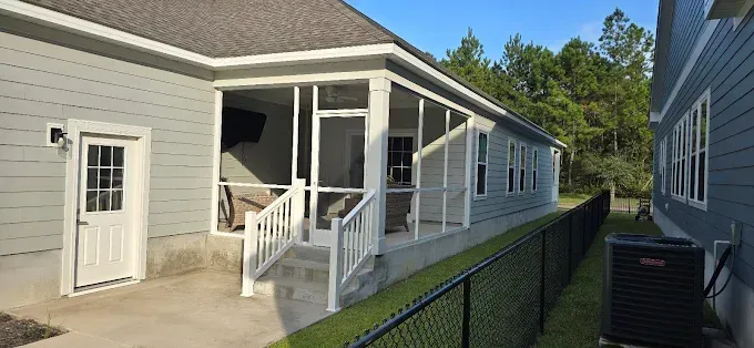 Exterior view of a screened porch attached to a light blue house, with a black fence and trees in the background.