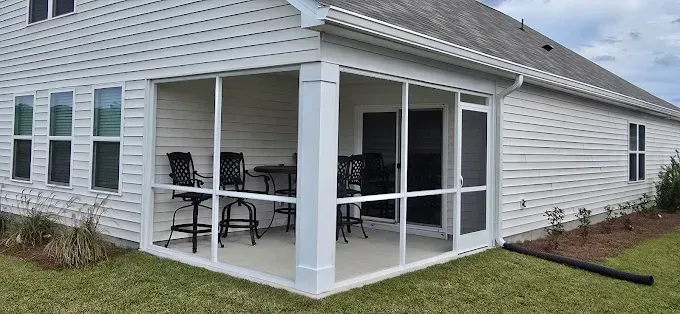 Screened-in porch on a white house, with chairs and table inside. Green grass and blue sky.