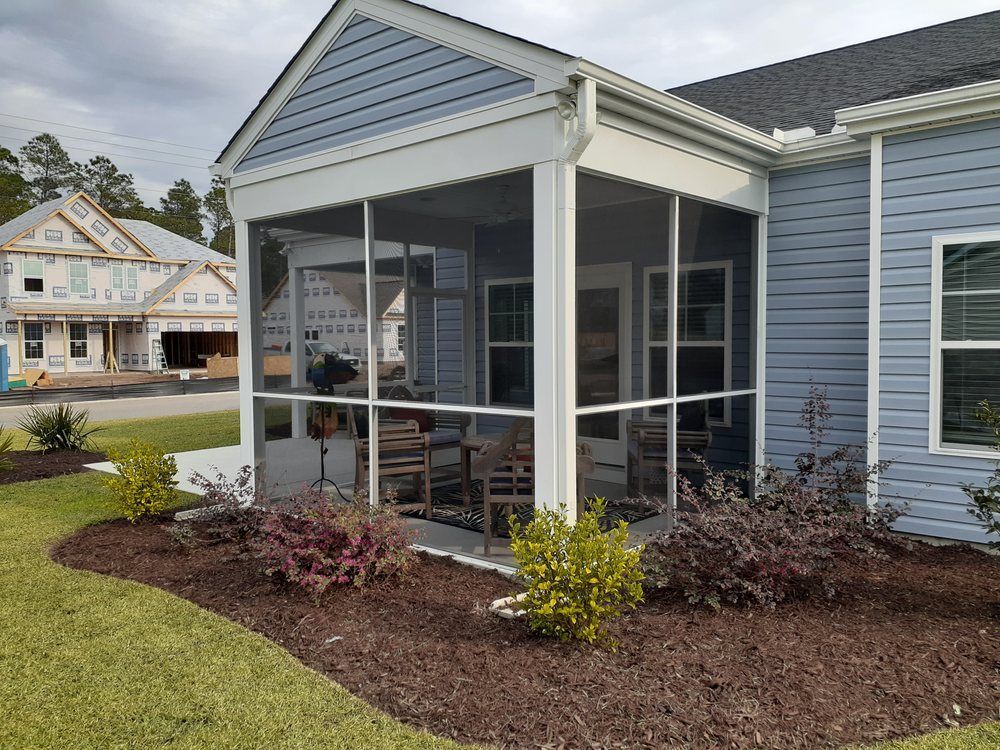 Screened-in porch on a blue house. Chairs and table visible inside, surrounded by landscaping and mulch.