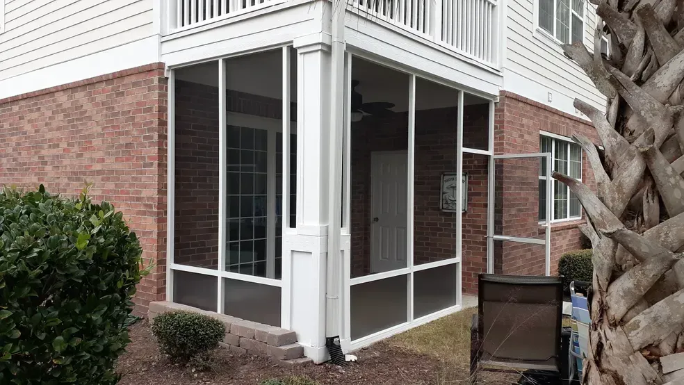 Screened-in porch on a brick building corner. White frames, beige flooring. A single door leads inside.