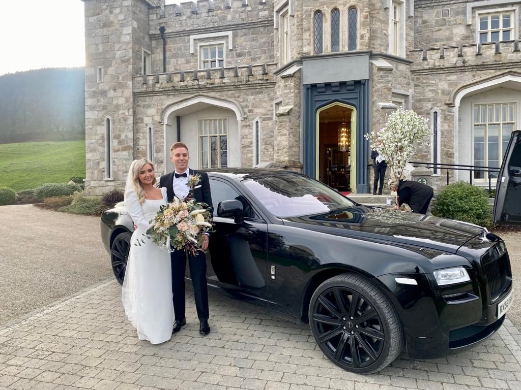 Wedding couple posing beside a black luxury car outside a stone building