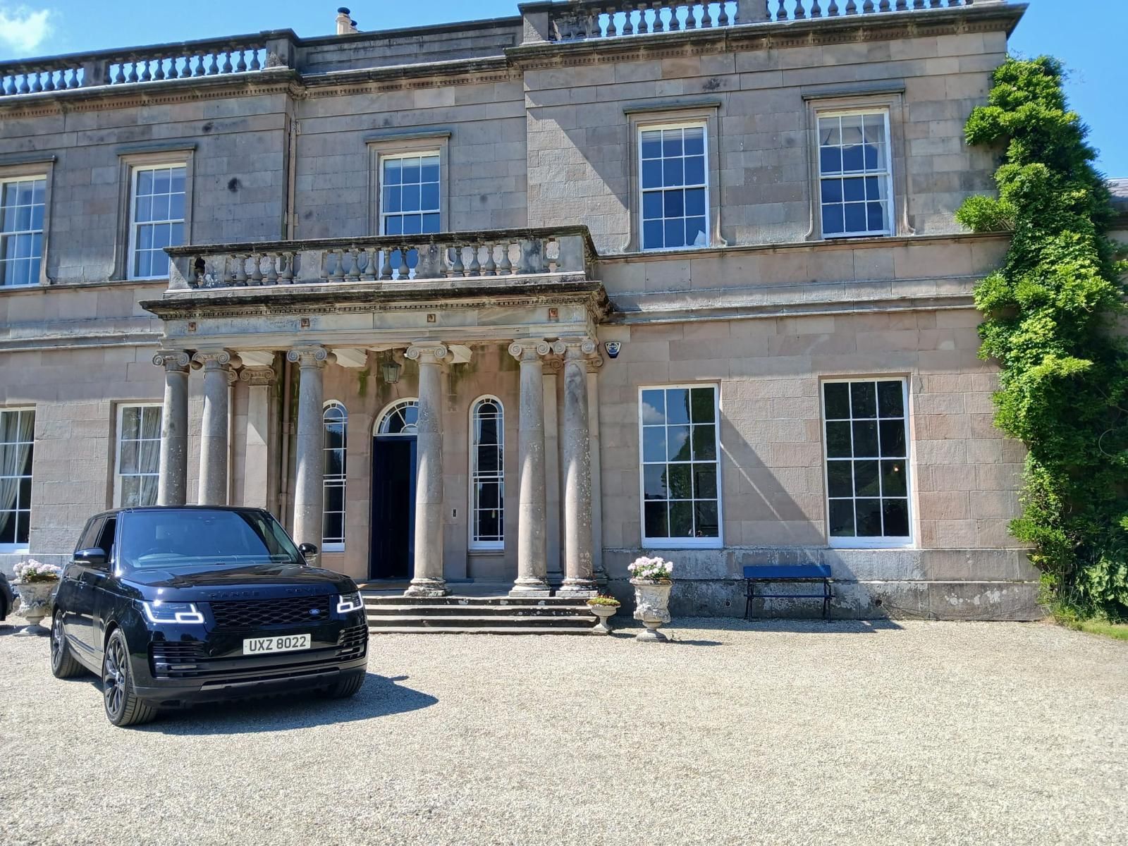 Grand stone mansion with columns, black car parked in front, and ivy on the right side