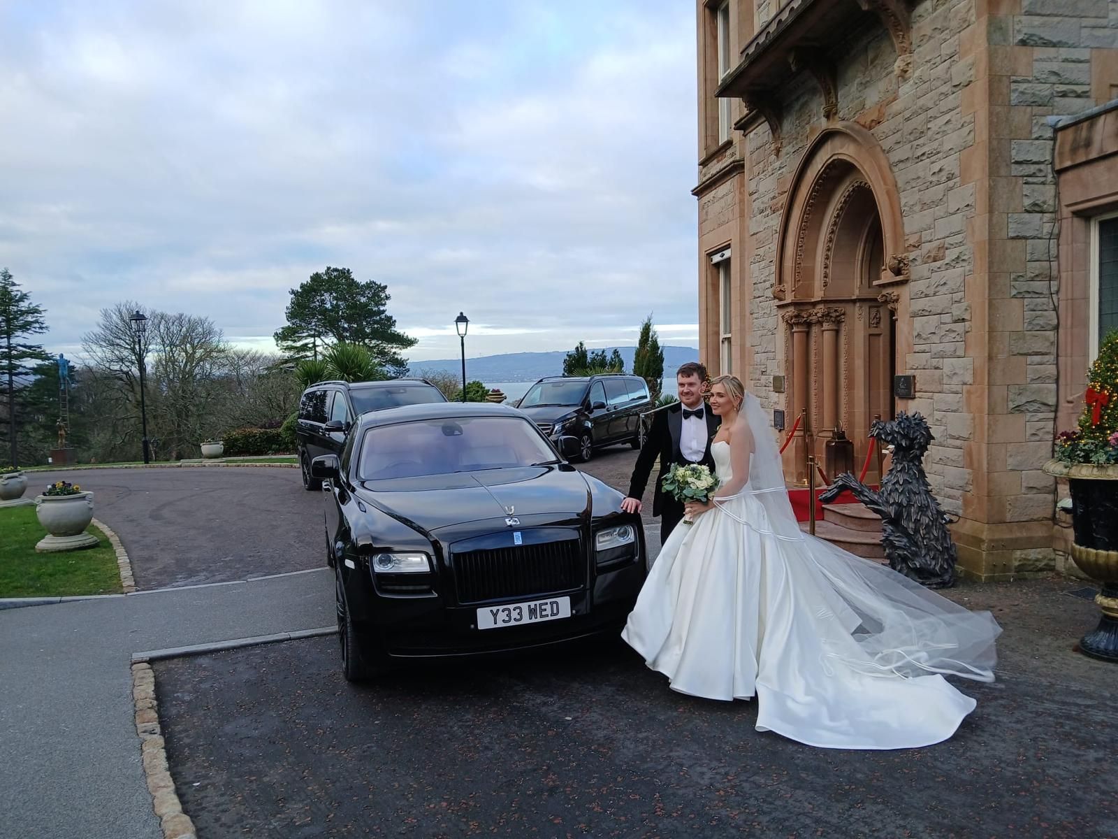 Bride and groom beside a black car outside a stone building with an arched doorway