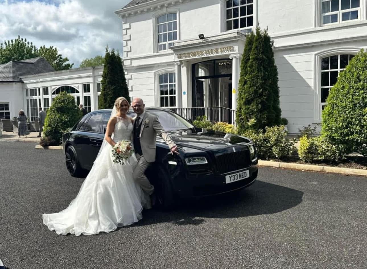 Bride and groom posing beside a black car in front of a white house