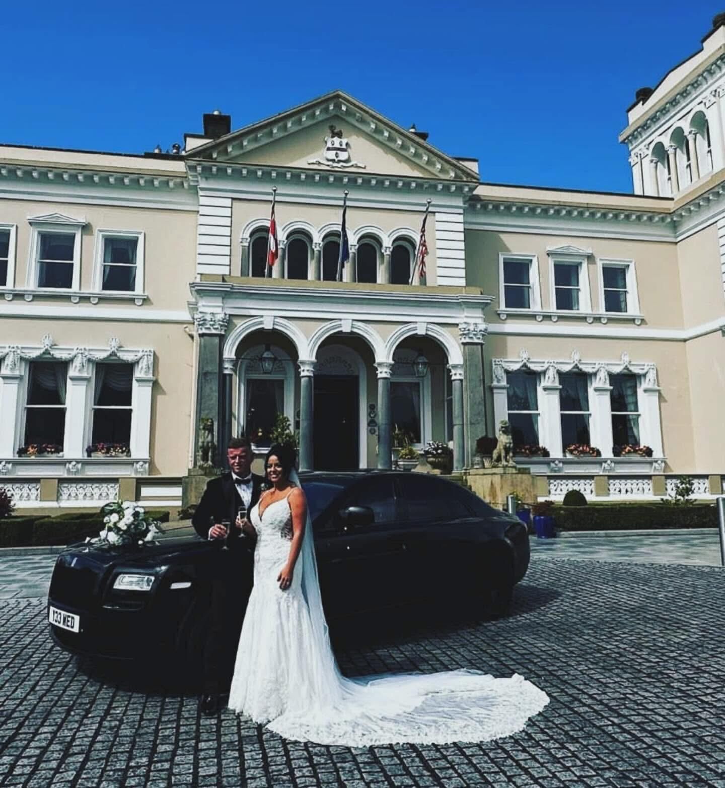 Bride and groom pose beside a black car in front of an ornate historic building.