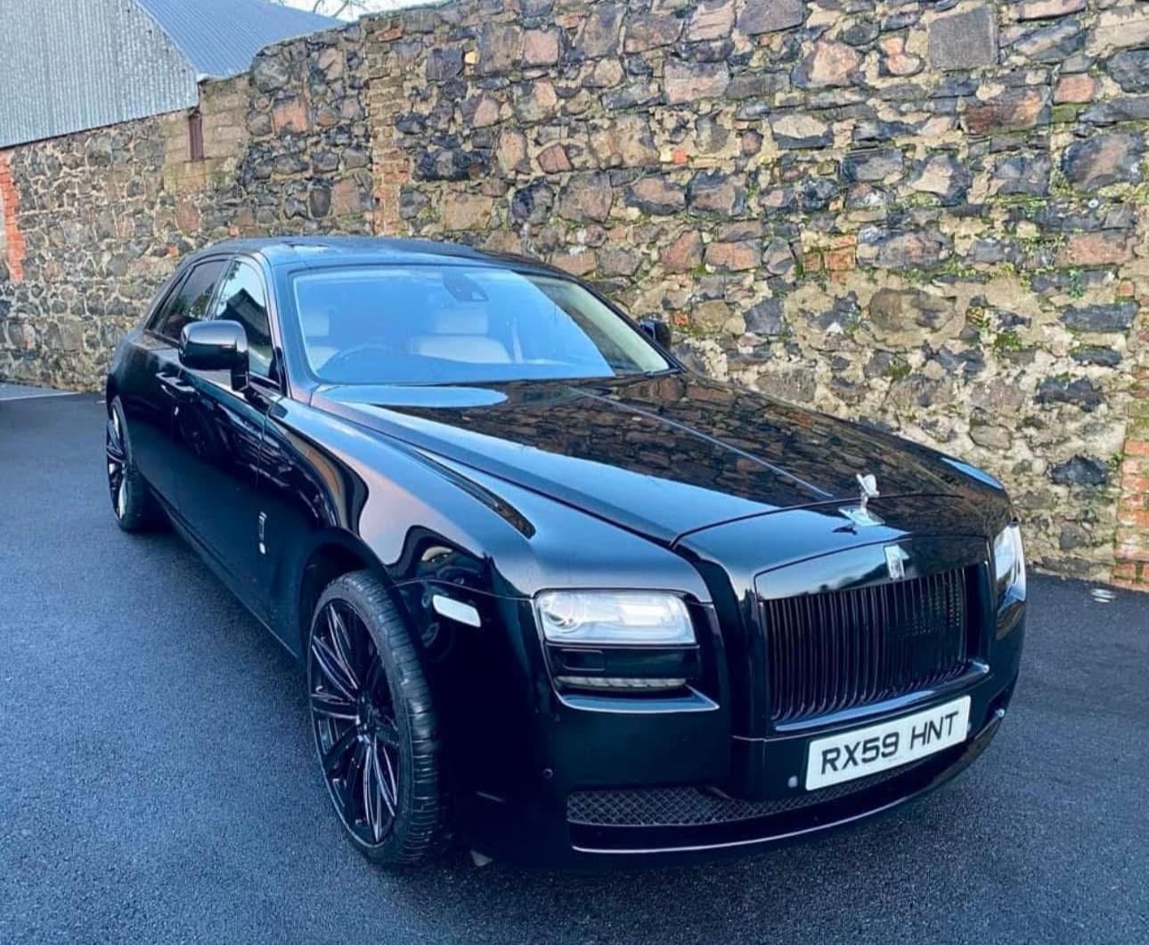 Black Rolls-Royce sedan parked on a wet road beside a stone wall