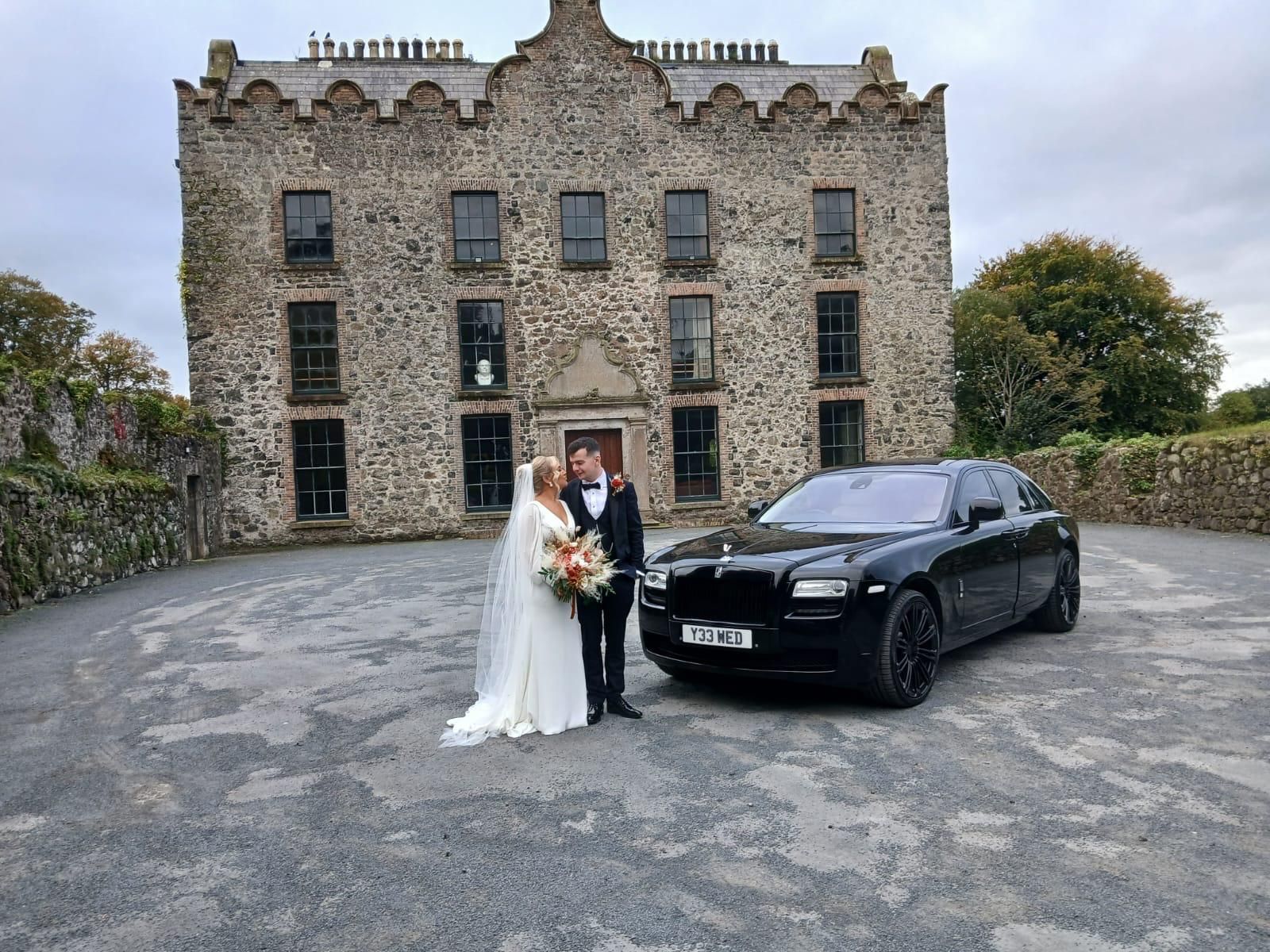 Bride and groom pose beside a black luxury car in front of a stone manor house.