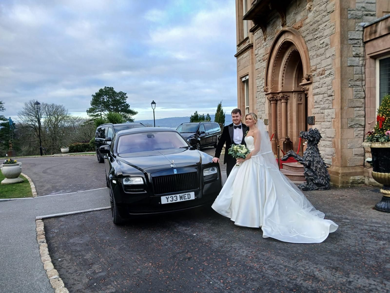 Bride and groom posing beside a black car outside a stone building