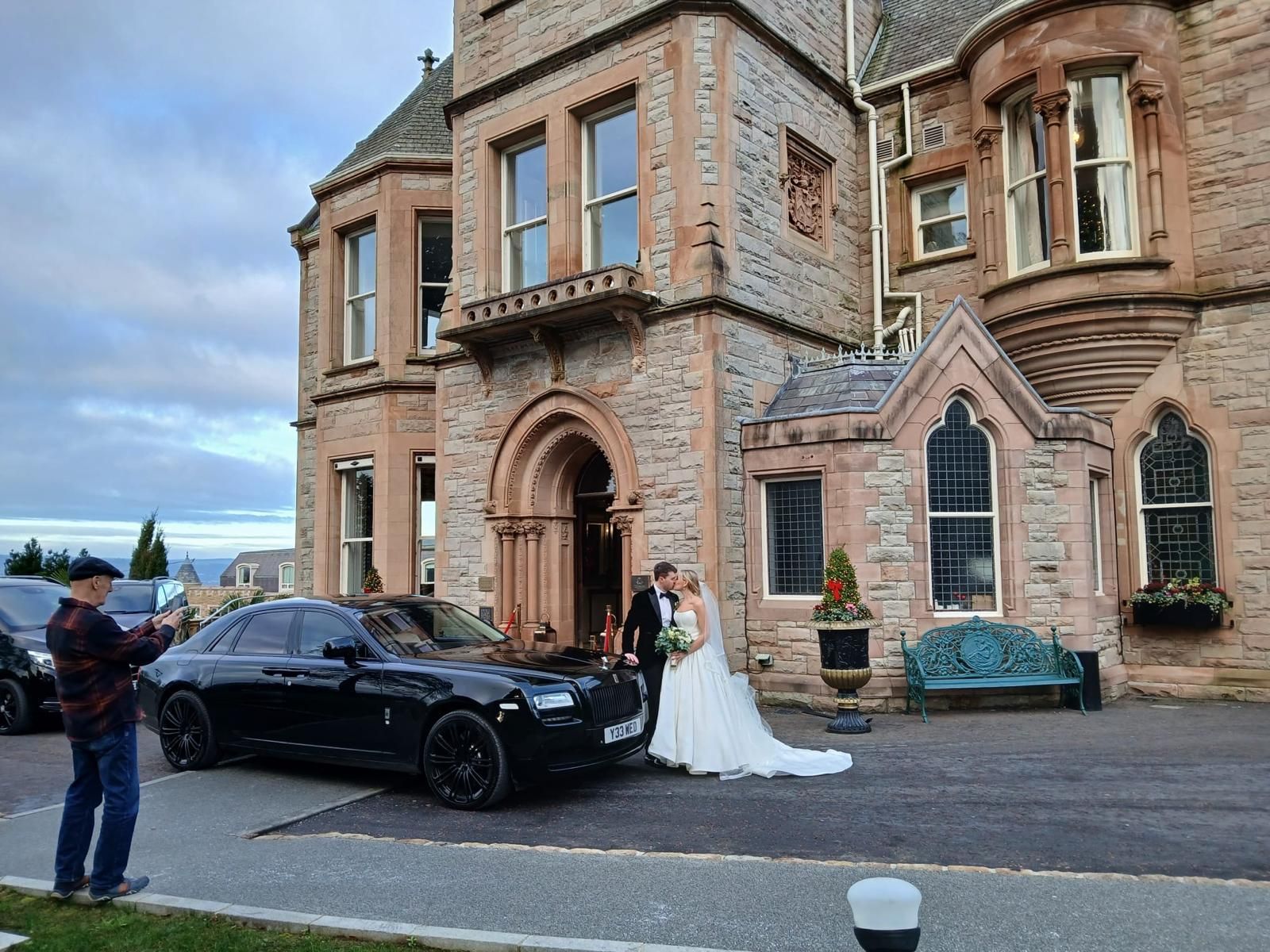 Bride and groom beside a black sports car outside a stone manor house