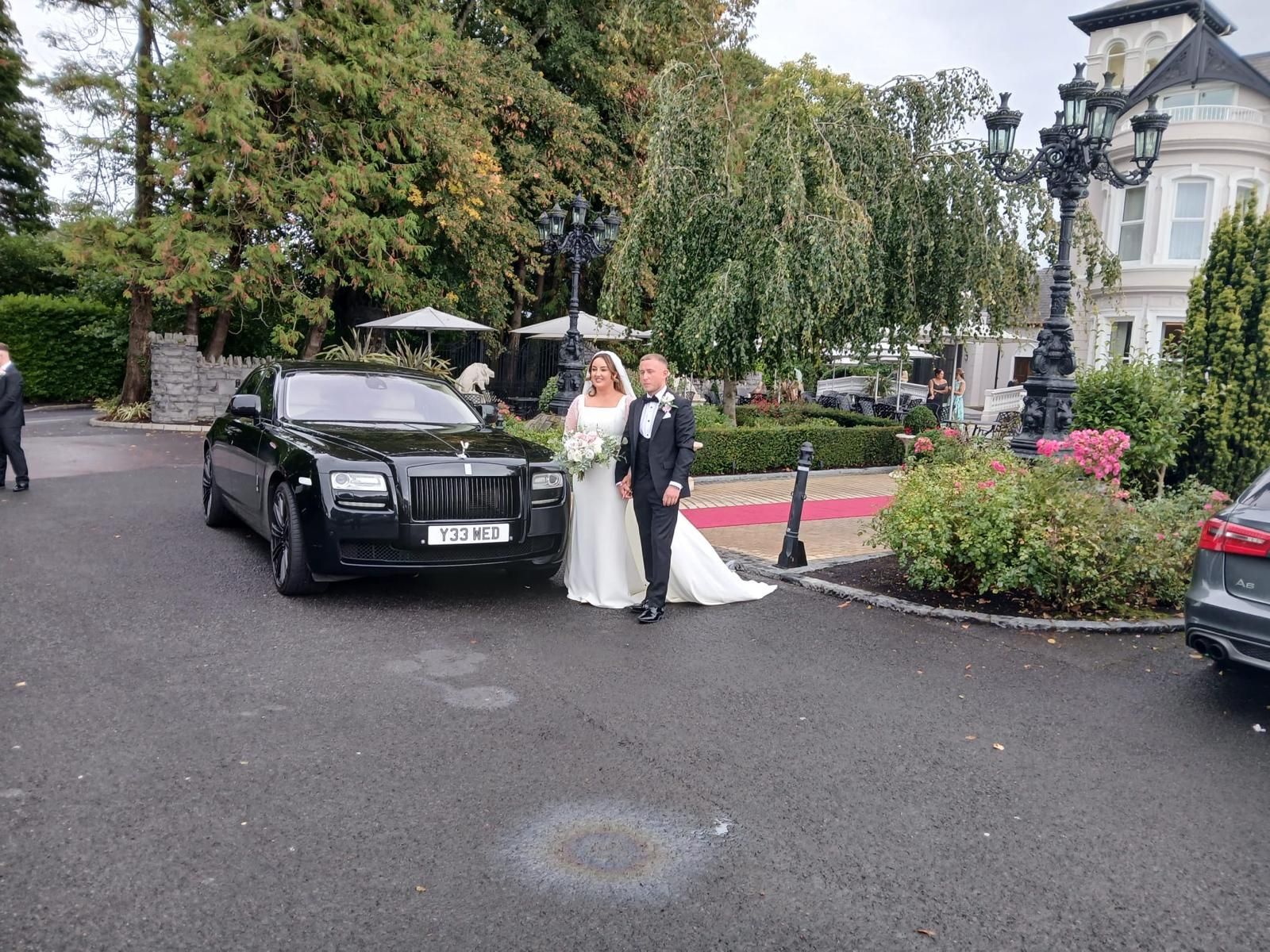 Bride and groom posing beside a black limousine in a garden driveway