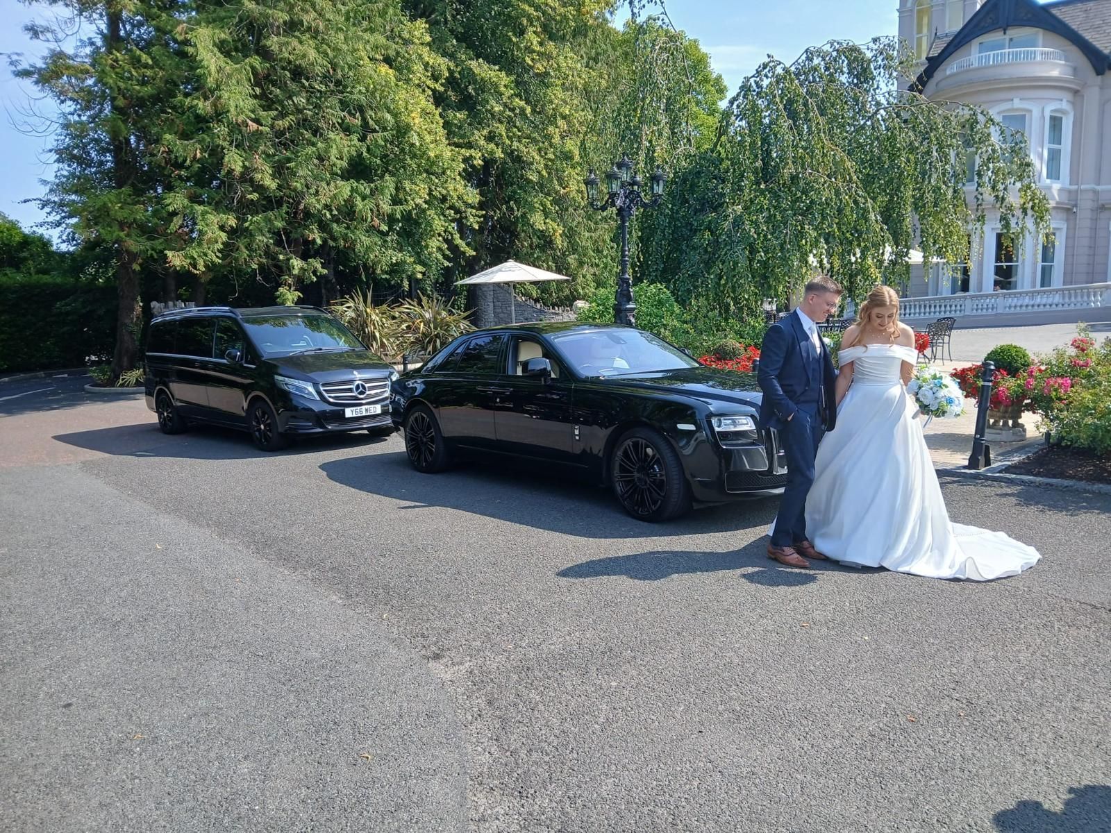Bride and groom with two black cars outside a white building on a sunny day