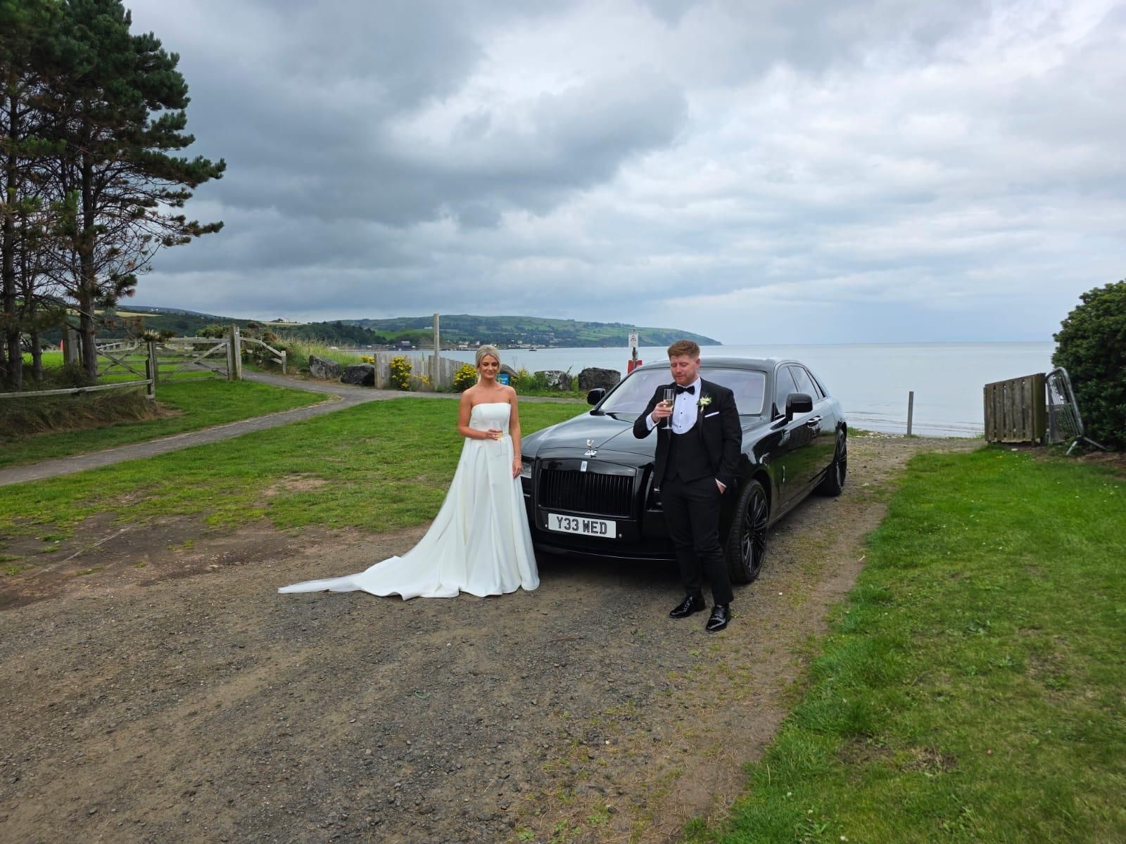 Wedding couple posing beside black limousine on a gravel path near the water