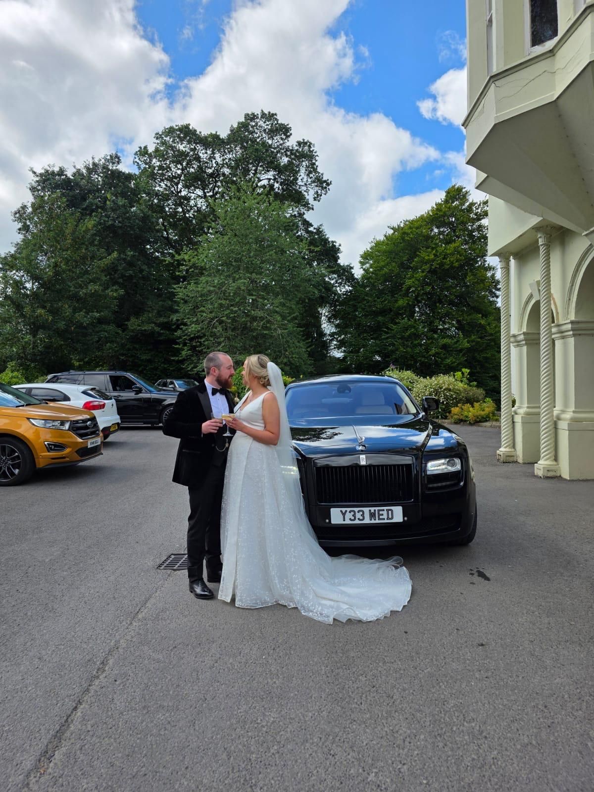 Bride and groom posing beside a black car outside a large building