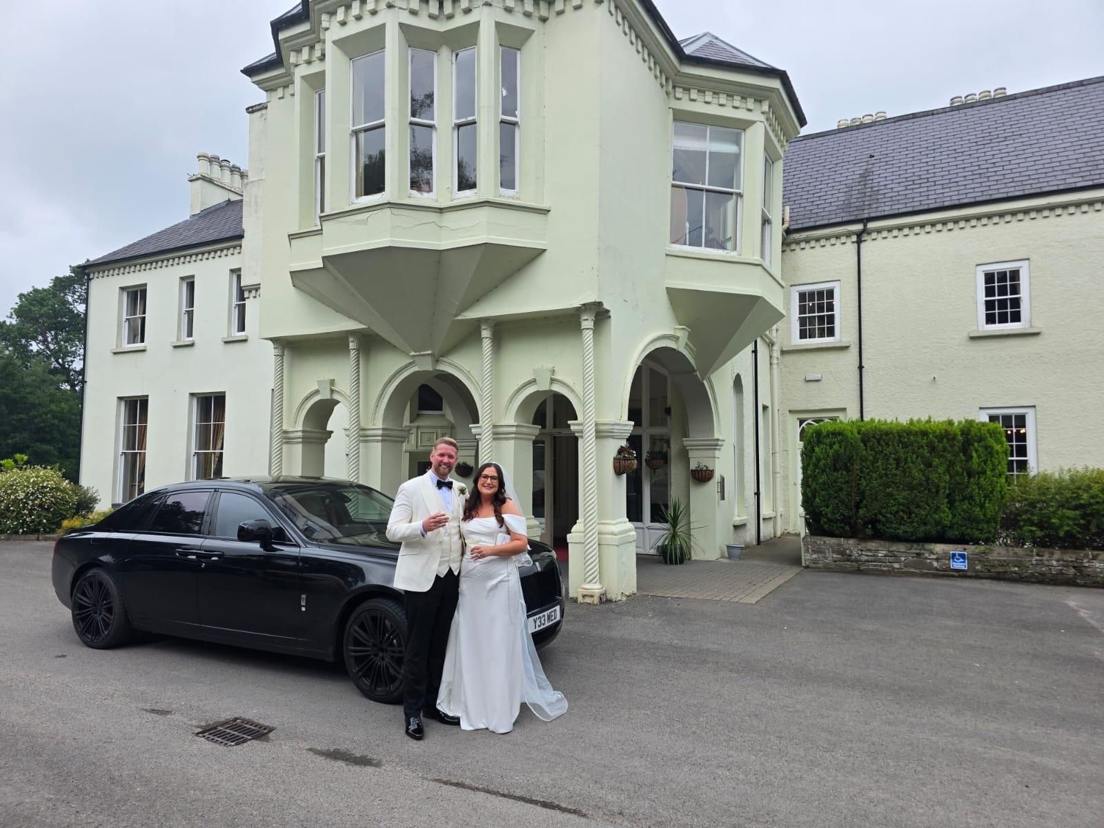 Newlyweds posing beside a black car outside a pale green manor house