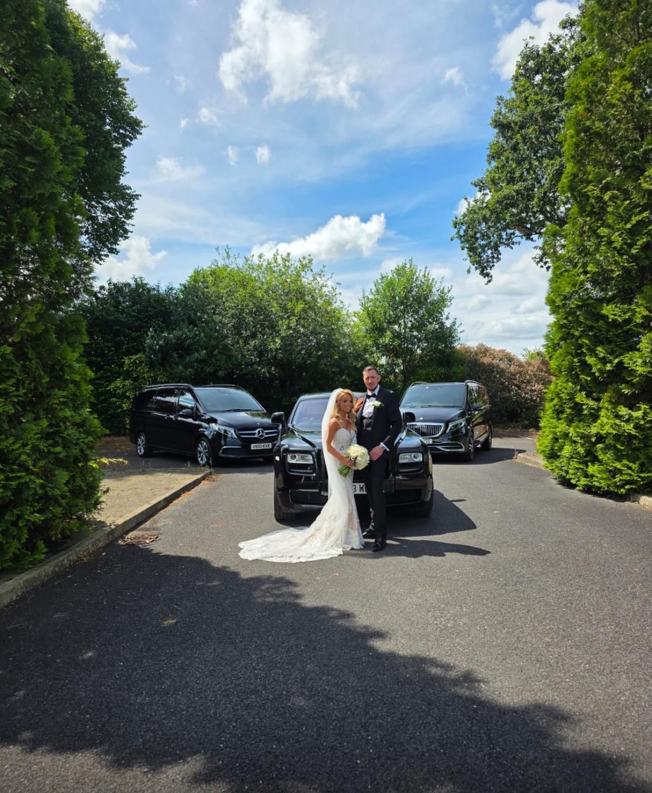 Bride and groom posing with cars on a driveway in bright sunlight