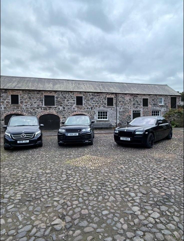 Three parked cars in front of a stone building on a cobblestone courtyard under cloudy skies.