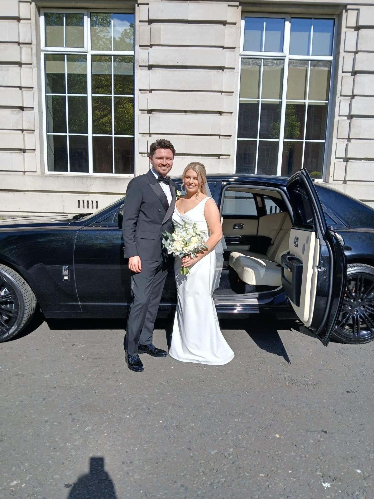 Bride and groom stand beside a black car with the door open outside a stone building.