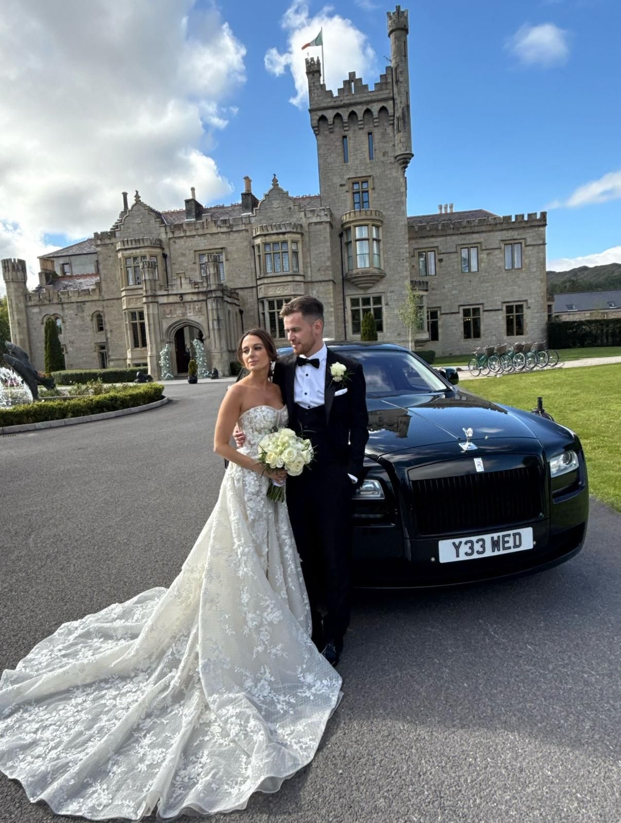 Bride and groom posing beside a black car in front of a castle-like mansion
