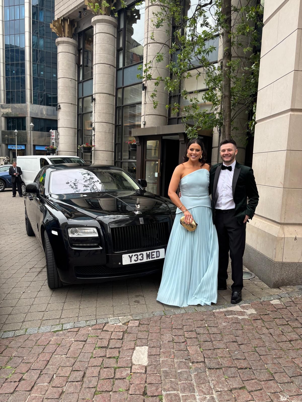 Couple posing beside a black car outside a modern building, with the woman in a light blue gown.
