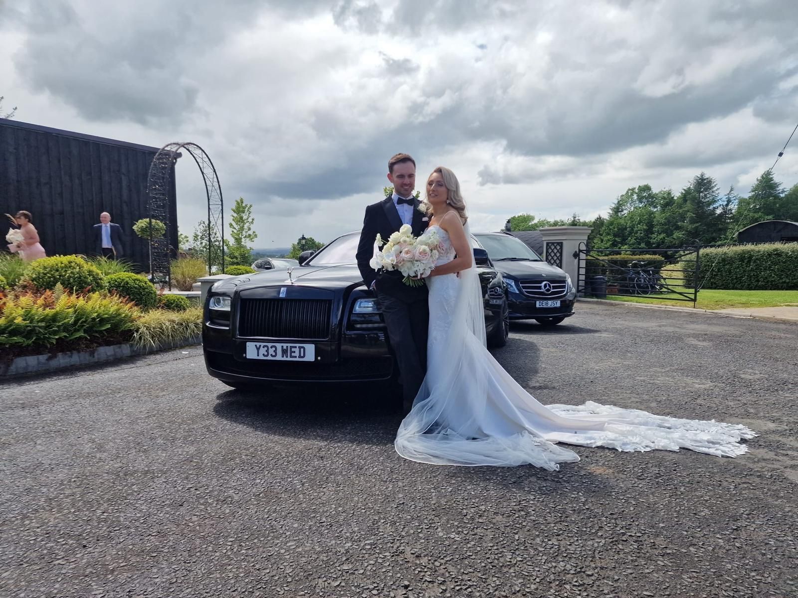 Bride and groom posing by a black car in a gravel driveway under cloudy skies