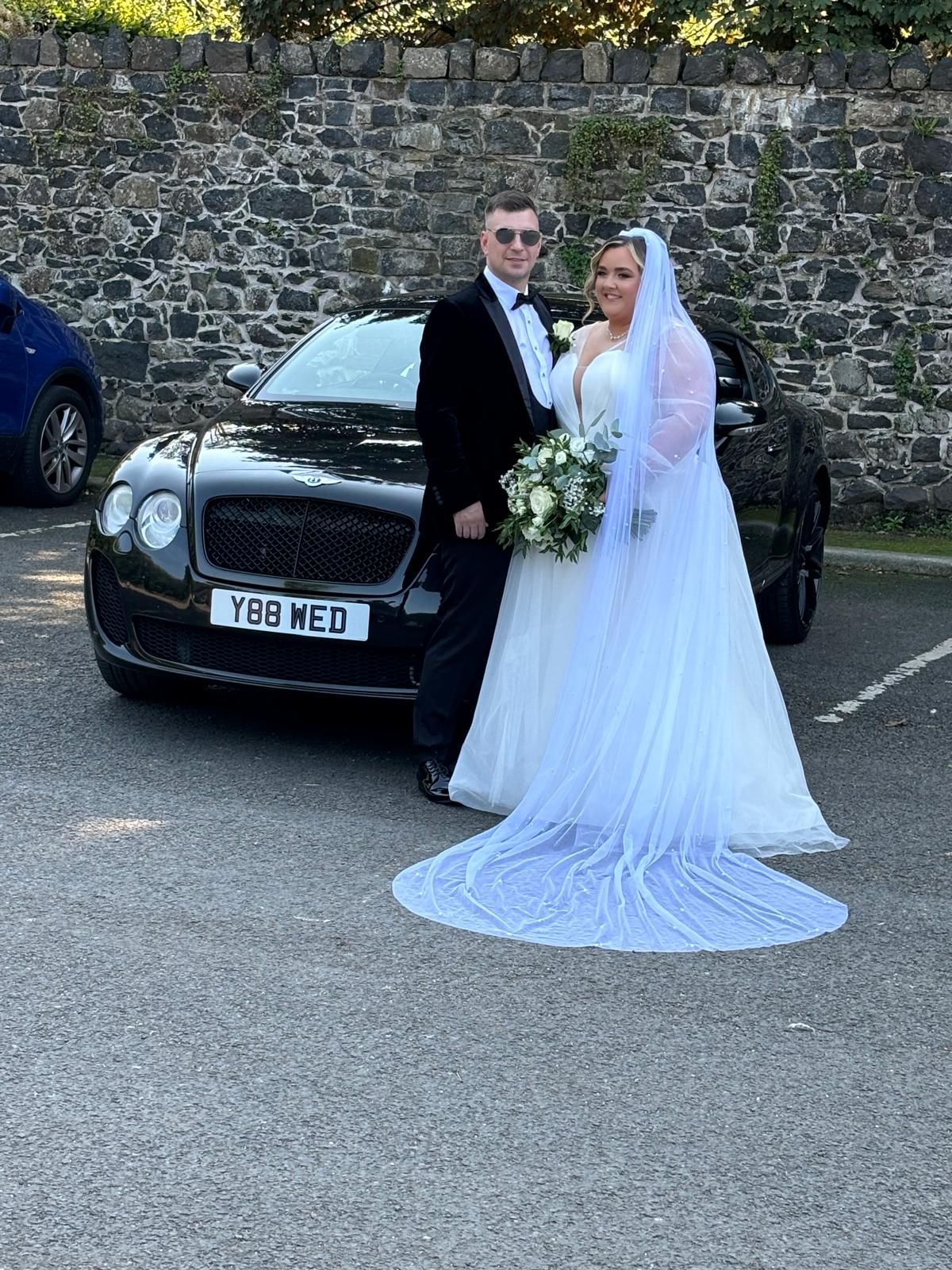 Bride and groom posing beside a black car in front of a stone wall outdoors