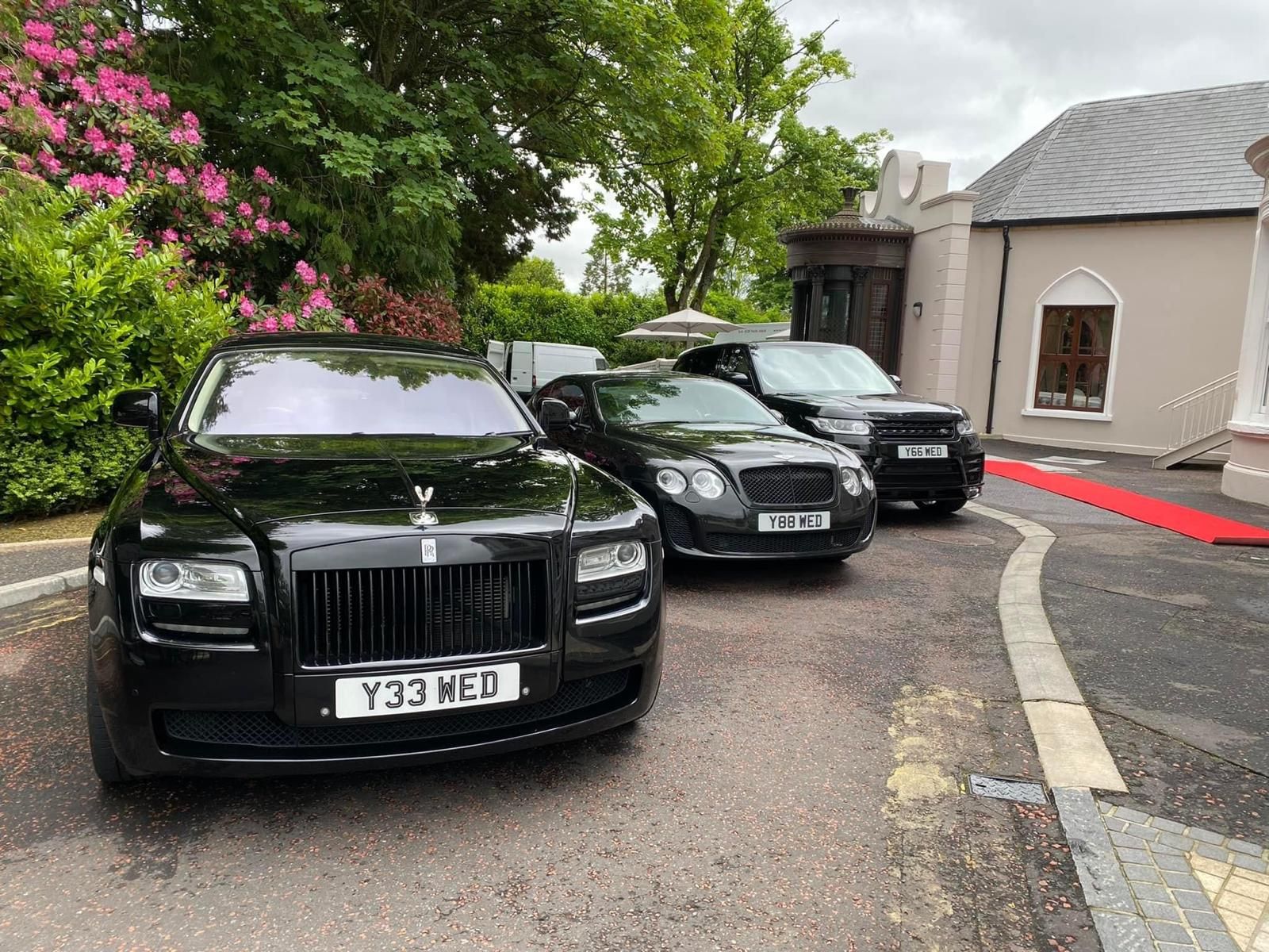 Two black Rolls-Royce cars parked beside a white building and red curb on a tree-lined drive