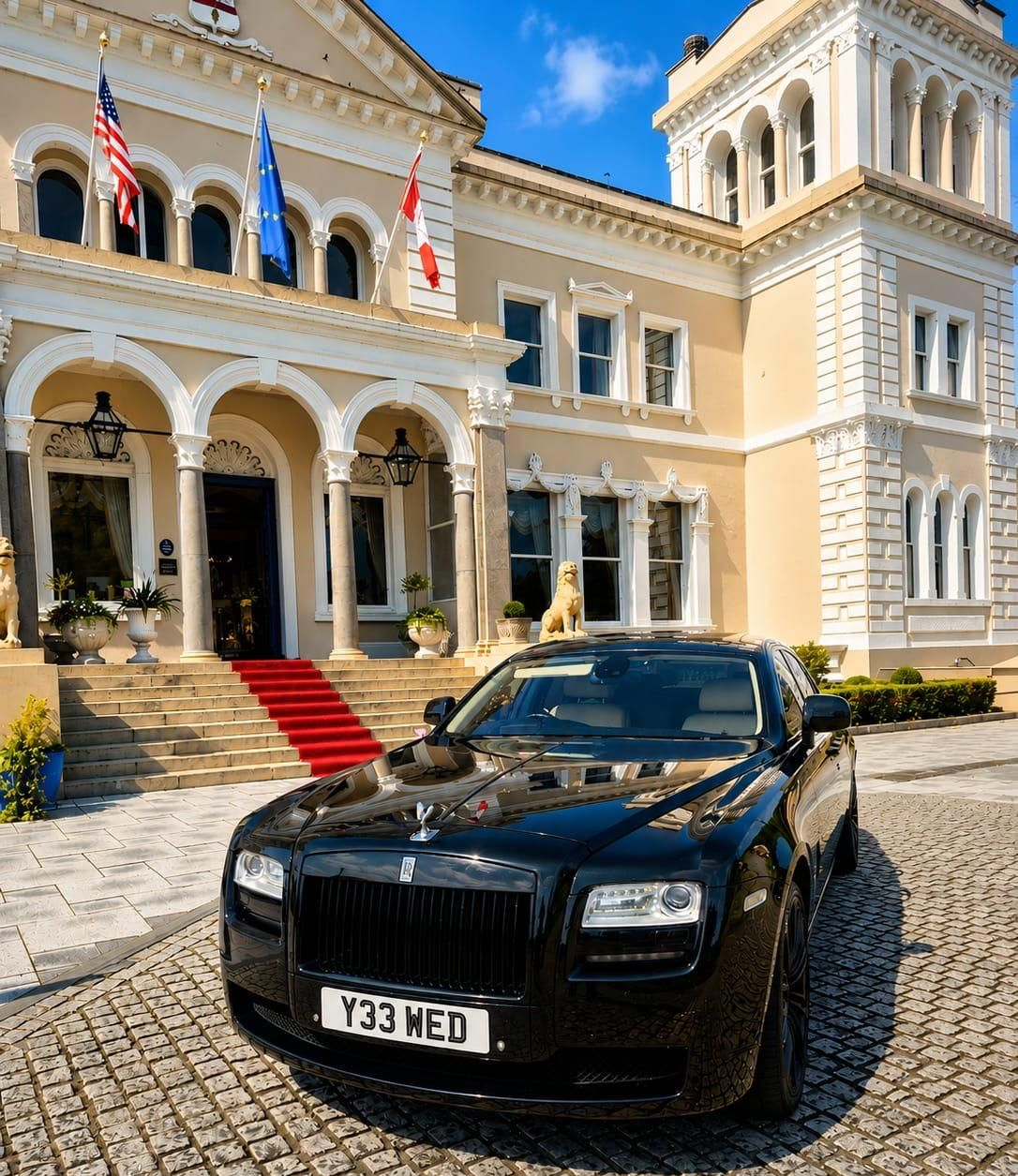 Black luxury car parked before a grand cream-colored mansion with red carpeted steps and flags.